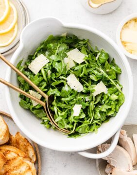 Two spoons resting in a bowl with arugula salad and shaved cheese.