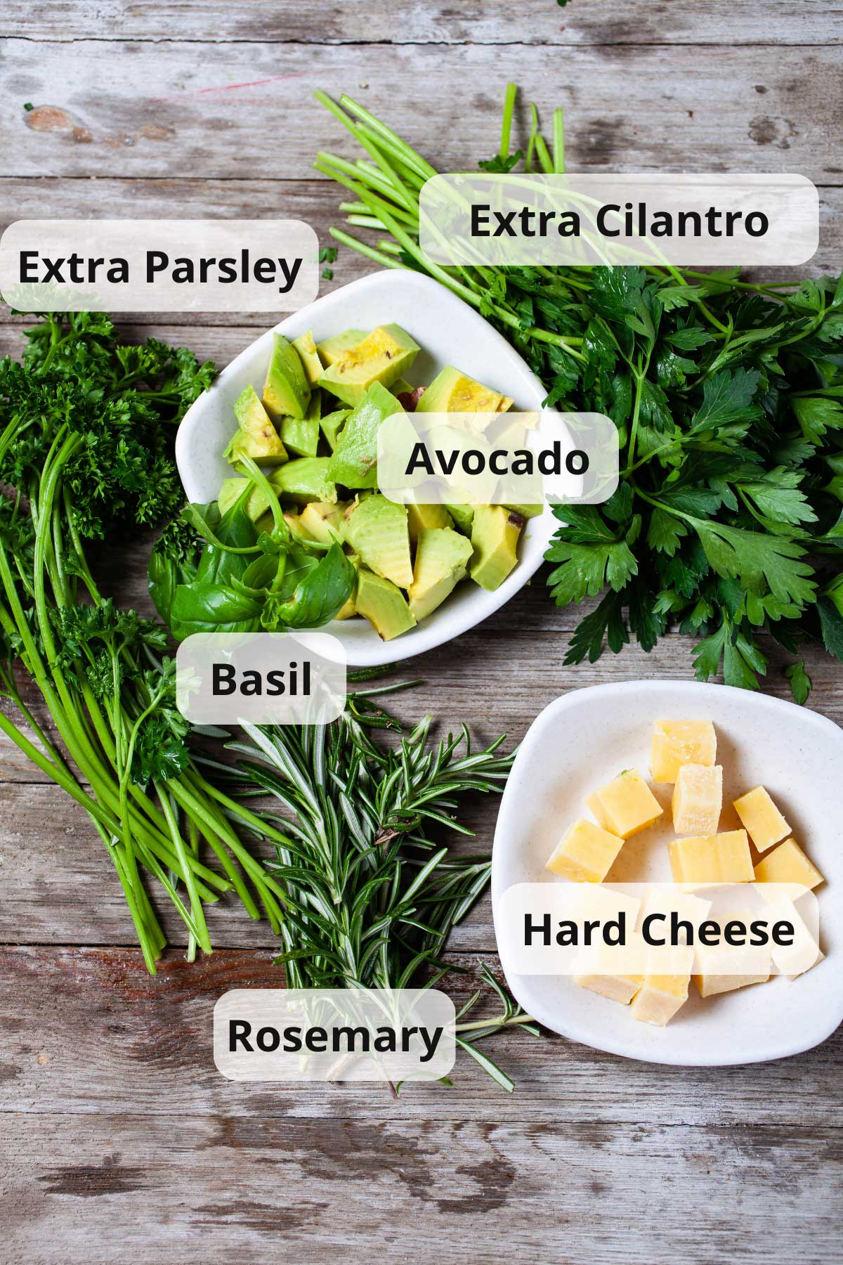 A wooden board with fresh ingredients such as parsley leaves, cubed avocado, cilantro leaves, fresh basil leaves, cubed hard cheese, and sprigs of Rosemary.