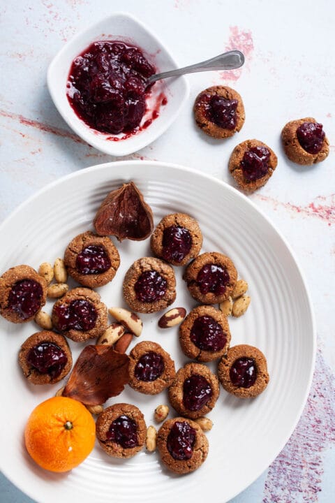 A plate filled with thumbprint jam cookies, nuts, and orange next to a bowl with jam.
