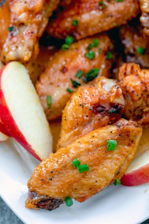 an up close shot of apple cider chicken wings on a white plate beside slices of fresh apples.
