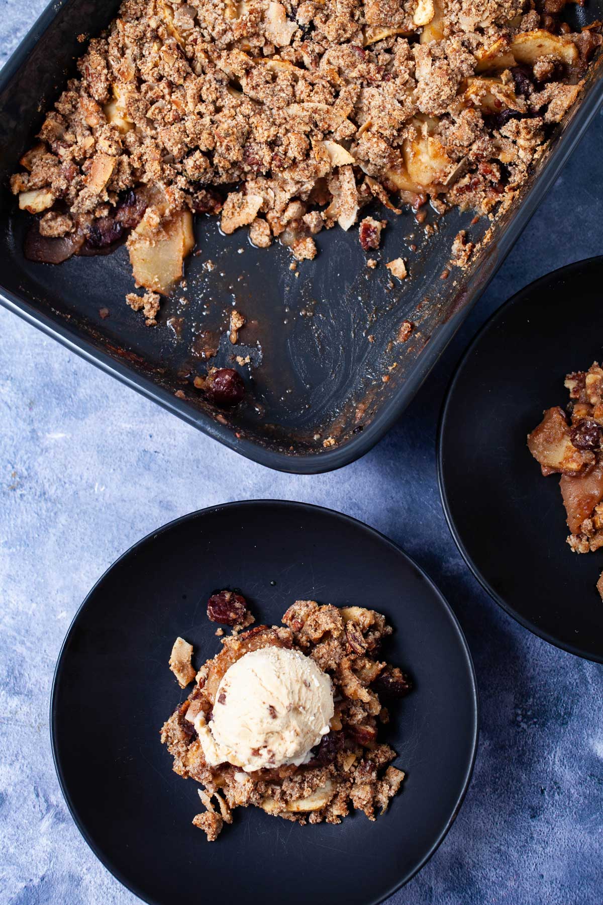 Healthy apple crumble served on a plate and topped with a scoop of ice cream next to a baking dish with the same apple crumble.