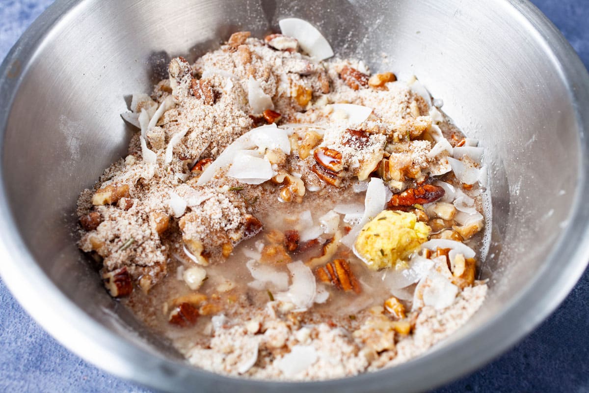 A dry flour mix with nuts and coconut shreds in a stainless steel bowl and topped with coconut oil and grated ginger.