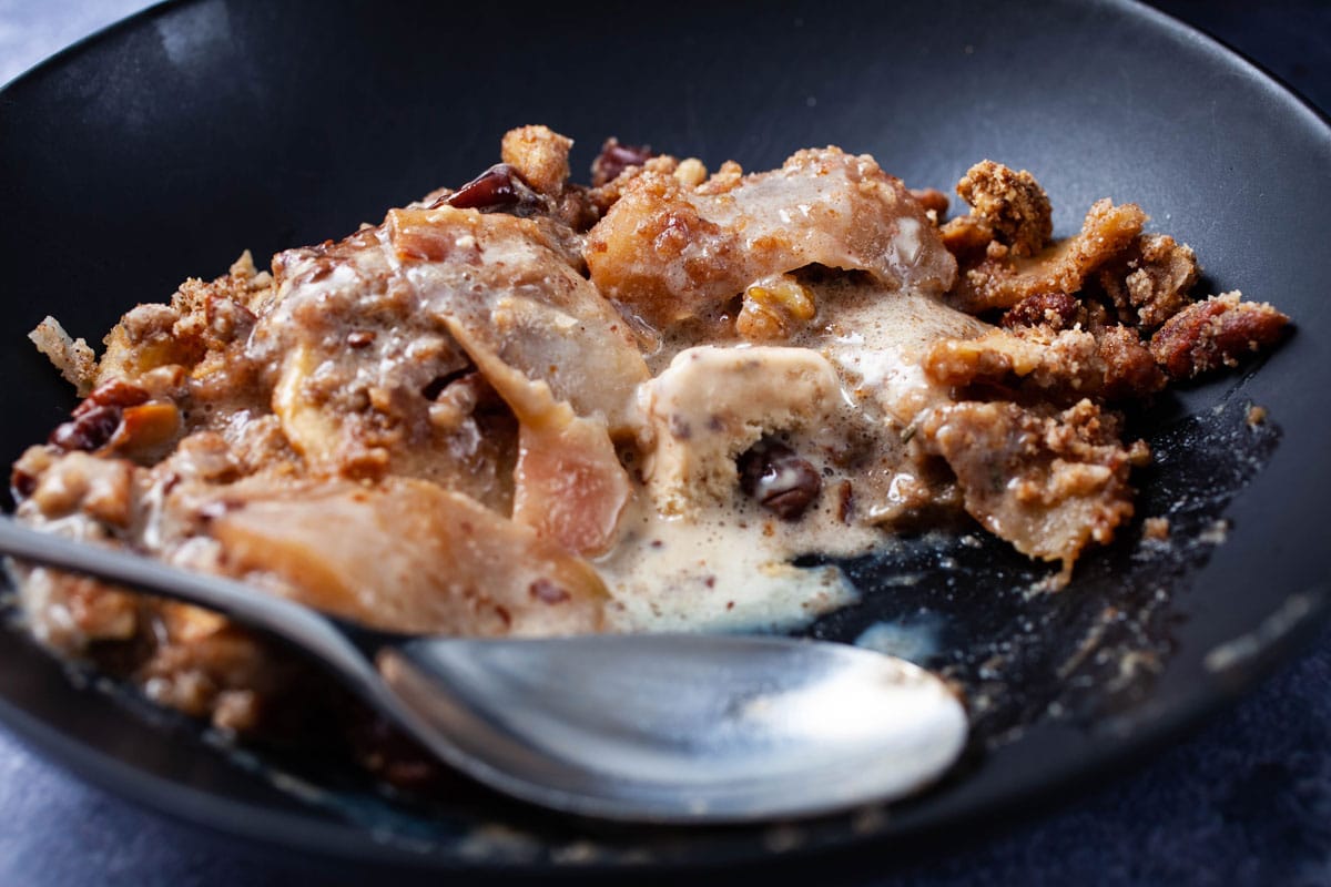 A close-up of a spoon resting in a plate with ice cream melted in a baked apple crisp.