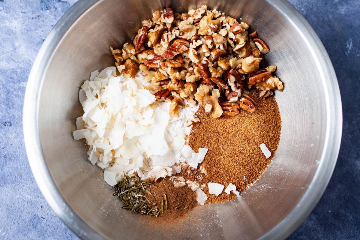 Dry ingredients like coconut shreds, herbs, nuts, coconut sugar in a stainless steel bowl.
