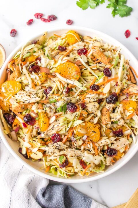 an overhead shot of an asian chicken salad in a large white bowl topped with dried cranberries, orange segments and poppy seeds.