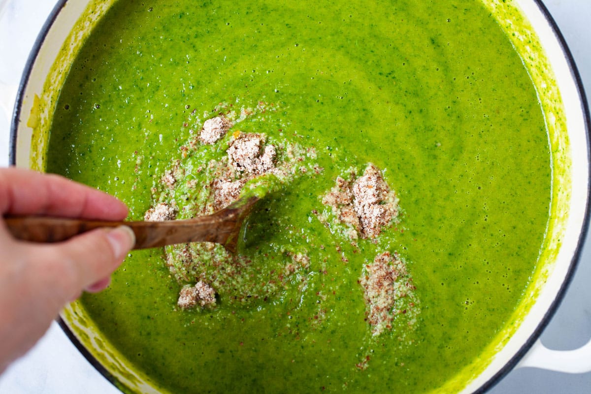 A hand stirring salt in creamy asparagus soup.