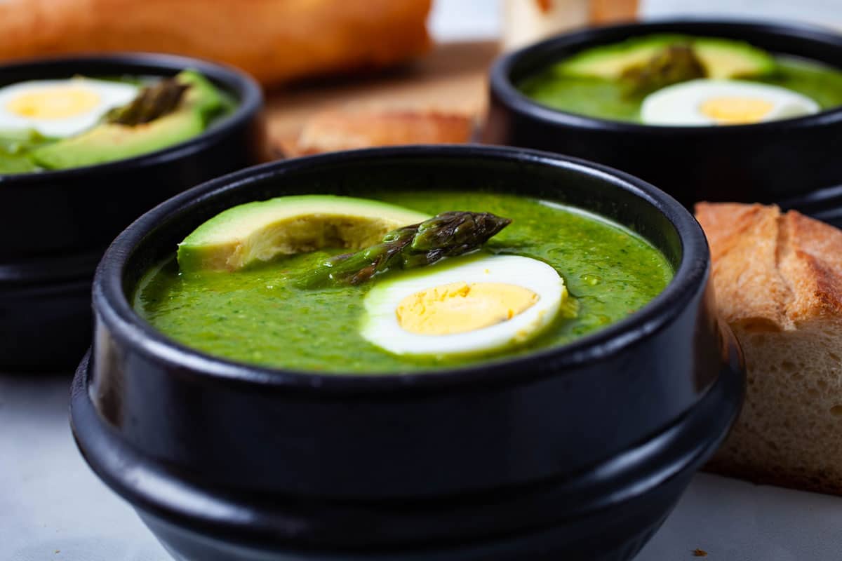 Three bowls of asparagus soup topped with avocado, half of a cooked egg and head of an asparagus with baguette bread on the side.