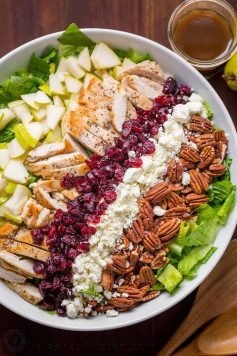 an overhead shot of a chopped chicken salad in a white bowl with the ingredients laid out on a bed of lettuce in neat rows, first pears, chicken, dried cranberries, cheese and pecans.