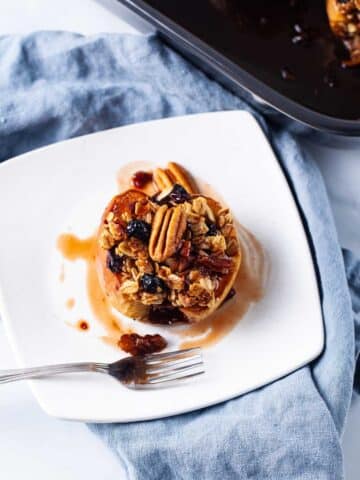 A small white plate filled with oatmeal stuffed baked apple and topped with raisins, nuts and syrup.