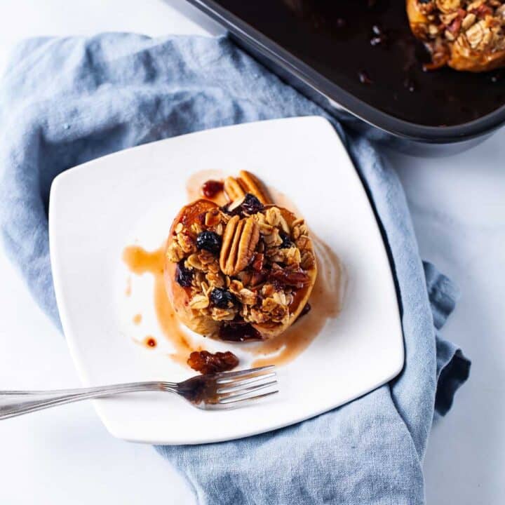 A small white plate filled with oatmeal stuffed baked apple and topped with raisins, nuts and syrup.