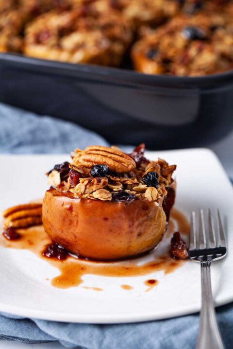 A small white plate filled with oatmeal stuffed baked apple and topped with raisins, nuts and syrup.