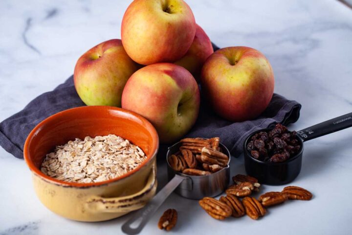 Red apples on a blue kitchen towel, a bowl of uncooked oatmeal, a fourth cup of dark raisins, and a third cup of half pecan nuts laid out on a table.