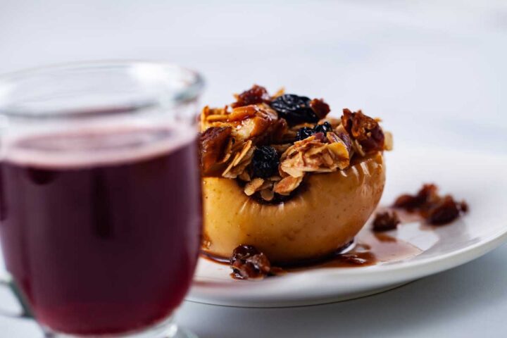 A close-up of a small glass filled with raspberry beer next to a stuffed and baked apple.