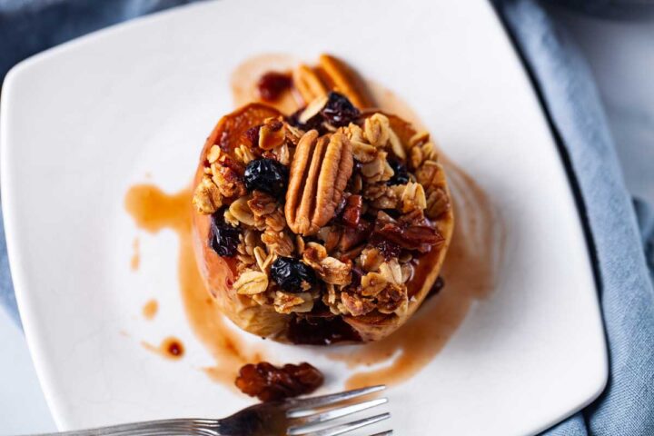 A small white plate filled with oatmeal stuffed baked apple and topped with raisins, nuts and syrup.