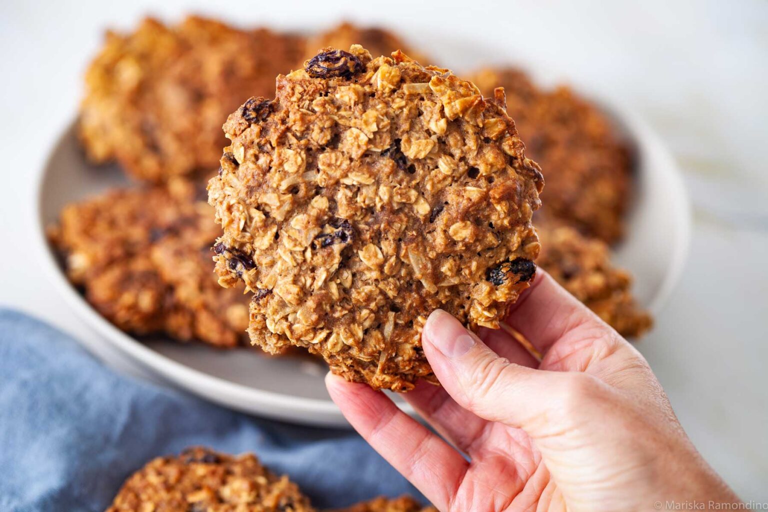 A hand holding an oat cookie with raisins, with a plate of more cookies in the background. The cookies appear freshly baked and golden brown. A blue cloth is partially visible to the side.