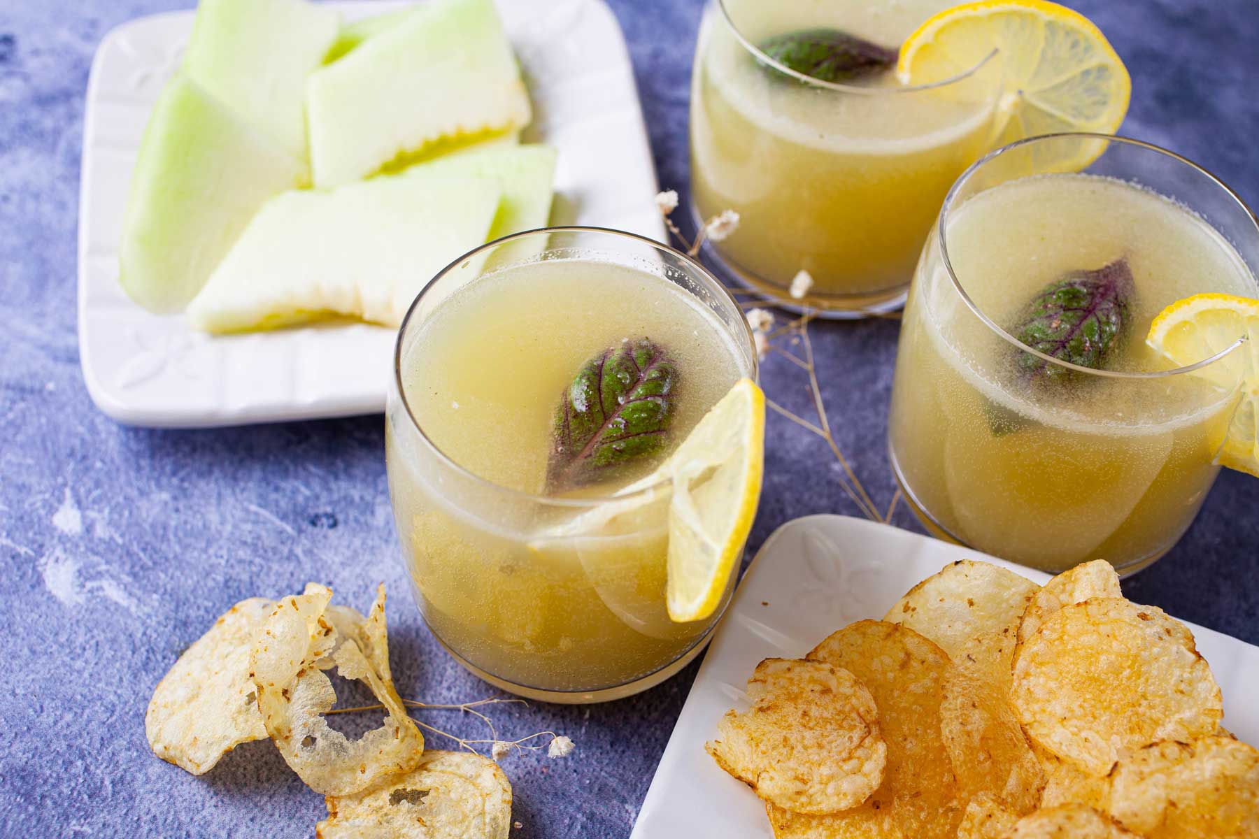 Fresh lemonade drinks garnished with basil leaves next to a plate with chips and sliced melon.