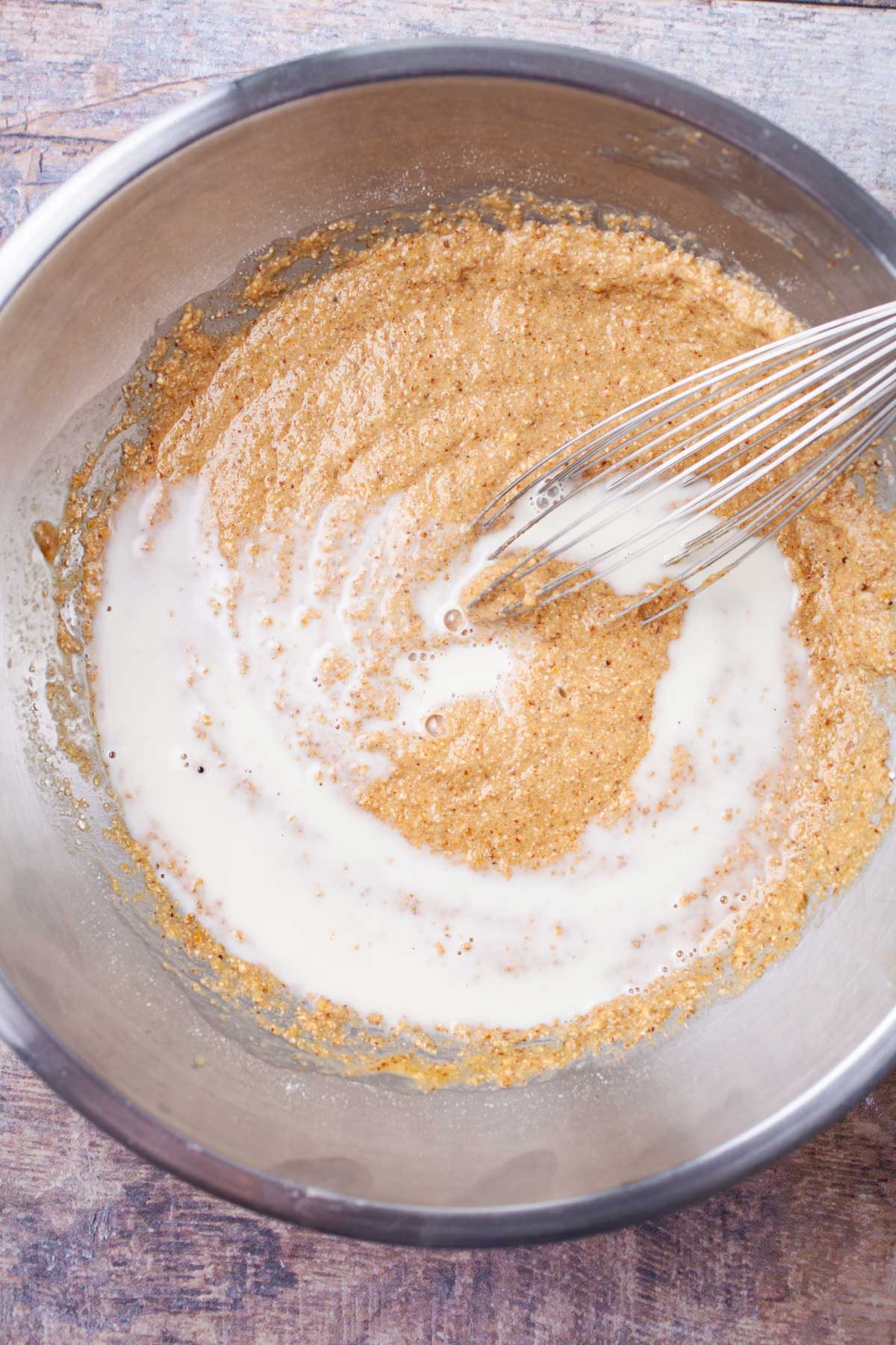 A whisk is combining milk with pancake batter in a stainless steel bowl.