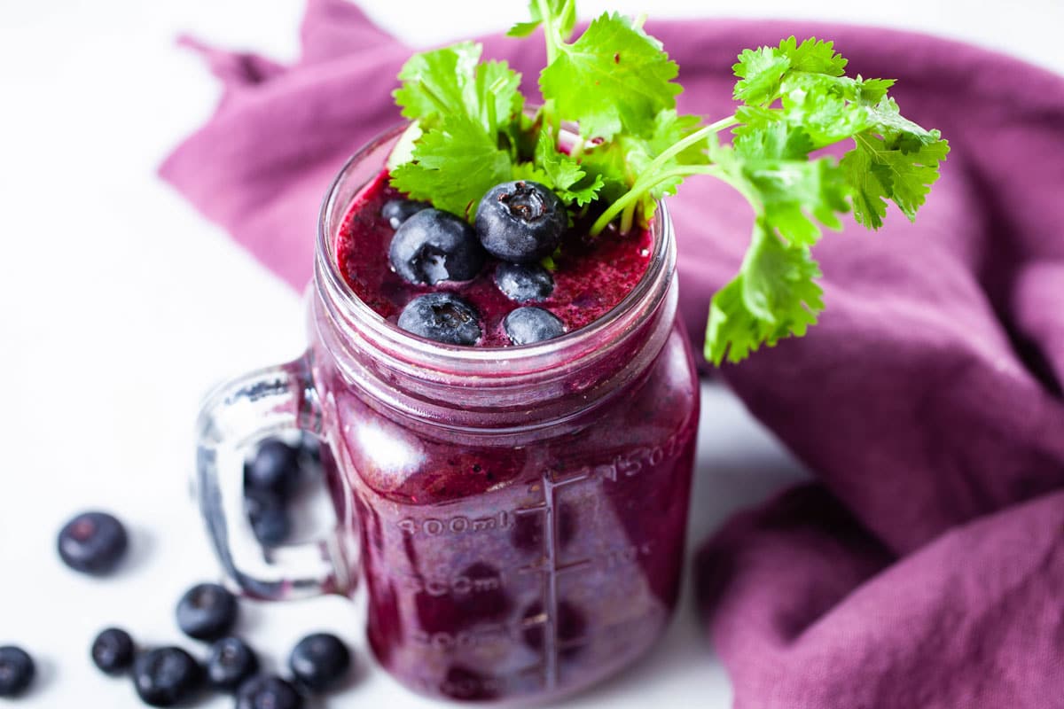 A glass of beet juice smoothie topped with blueberries and garnished with fresh cilantro leaves.