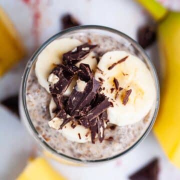 A cup full of banana chia pudding topped with fresh banana slices and shaved chocolate.