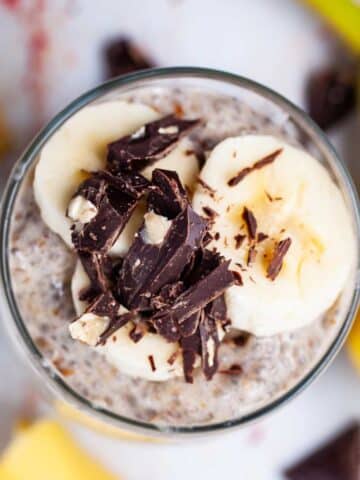 A cup full of banana chia pudding topped with fresh banana slices and shaved chocolate.