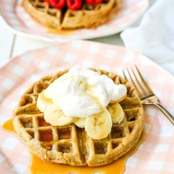 A round waffle on a plate topped with slices of banana and coconut whip cream.