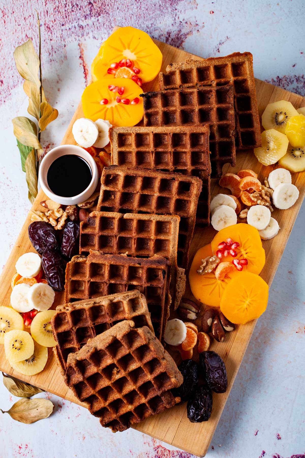 Golden brown waffles , an assortment of fresh and dried fruits, nuts, and a small syrup pourer beautifully arranged on a wooden board.