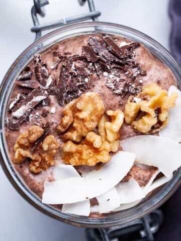 A chocolate chia pudding in a mason jar topped with coconut flakes, walnuts, and dark chocolate bits.