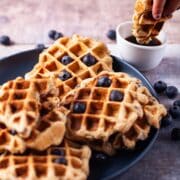 A hand dipping a piece of waffle in maple syrup next to a plate with oat flour waffles topped with blueberries.