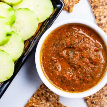 An Italian red pepper spread in a ramekin next to cucumber slices and toast.