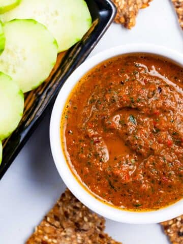 An Italian red pepper spread in a ramekin next to cucumber slices and toast.