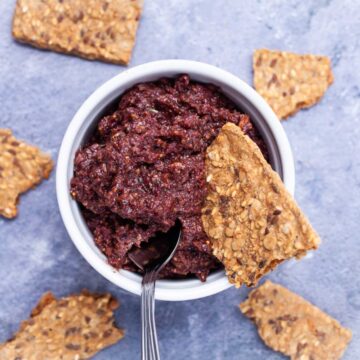 A spoon resting in a Kalamata olive and fig tapenade in a ramekin served with pieces of toast.