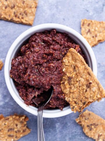 A spoon resting in a Kalamata olive and fig tapenade in a ramekin served with pieces of toast.