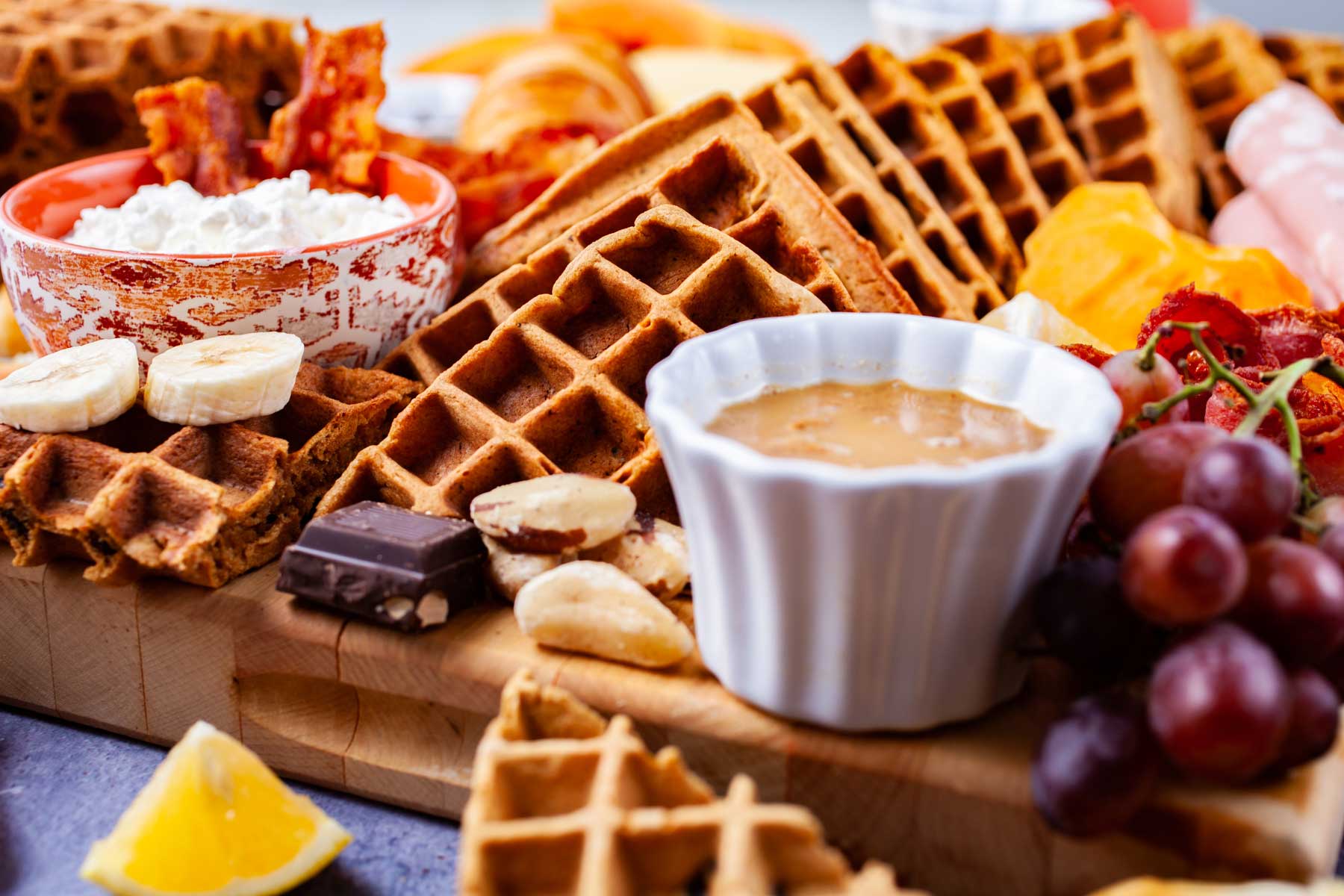 Fresh fruits, cheeses, charcuterie, meats, chocolate, croissants, spreads, coffee, juices, nuts, and homemade waffles all beautifully displayed on a waffle board and table.