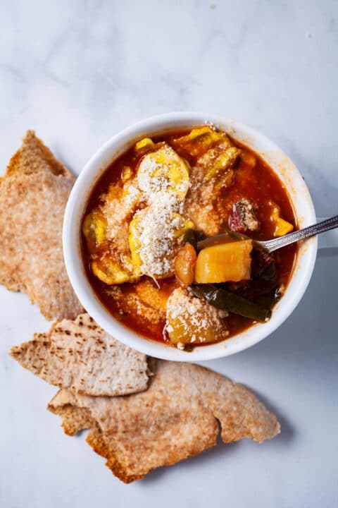 A cup of vegetable minestrone soup topped with plantain chips, grated cheese, and served with pitta bread on the side.