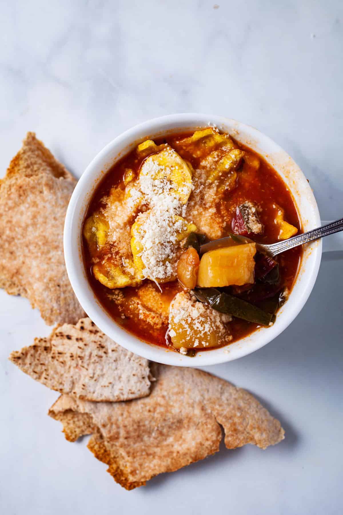 A spoon resting in a red tomato based vegetable soup topped with parmesan cheese.