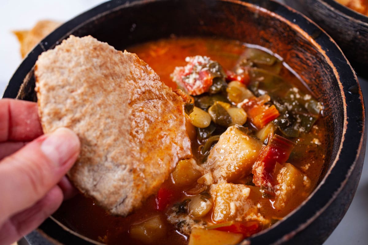 A hand dipping a piece of bread in a chunky vegetable soup.