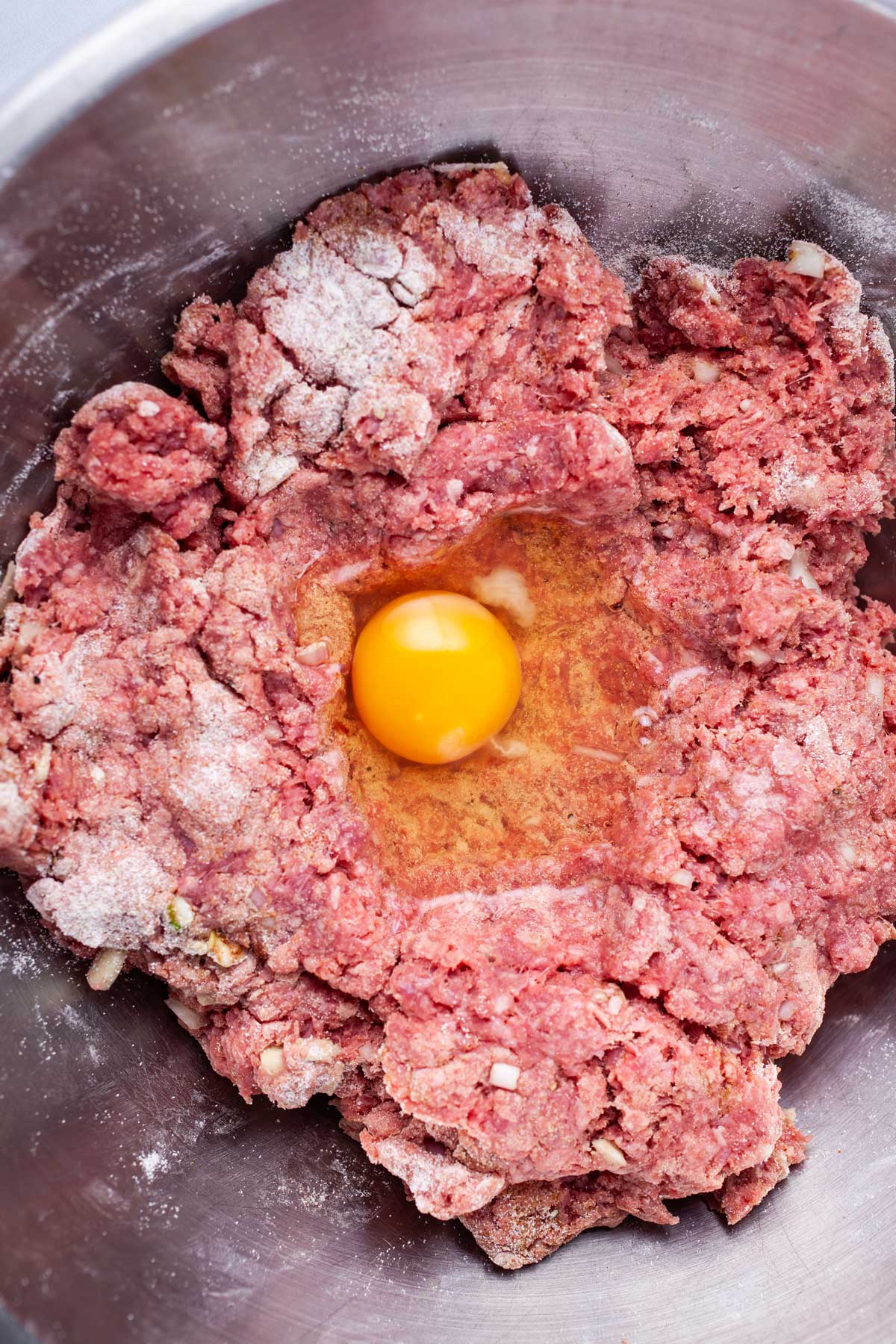 A raw egg nestled in a bed of raw meat in a stainless steel bowl.