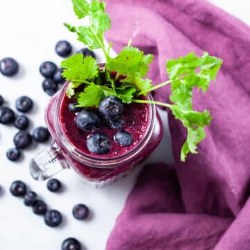 A glass of beet juice smoothie topped with blueberries and garnished with fresh cilantro leaves.