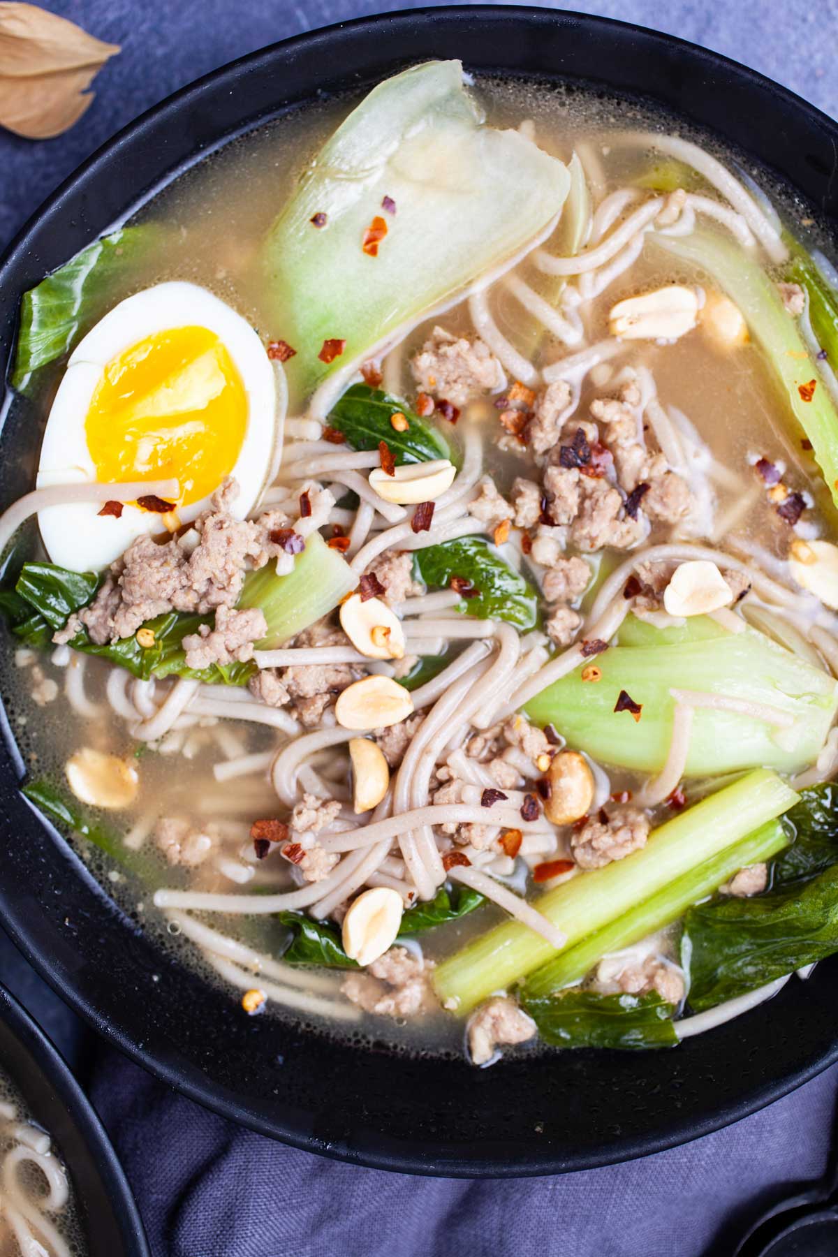 Bock choy soup with pork and noodles and garnished with chili peppers, peanuts, and hard-boiled egg.