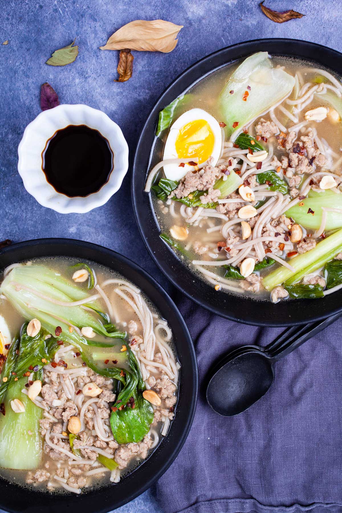 Two plates of bock choy soup with pork and noodles and garnished with chili peppers, peanuts, and hard-boiled egg.