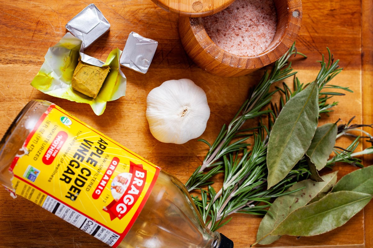 Apple cider vinegar, garlic, sea salt, Rosemary sprigs, Bay leaves, and bouillon cubes on a wooden table.