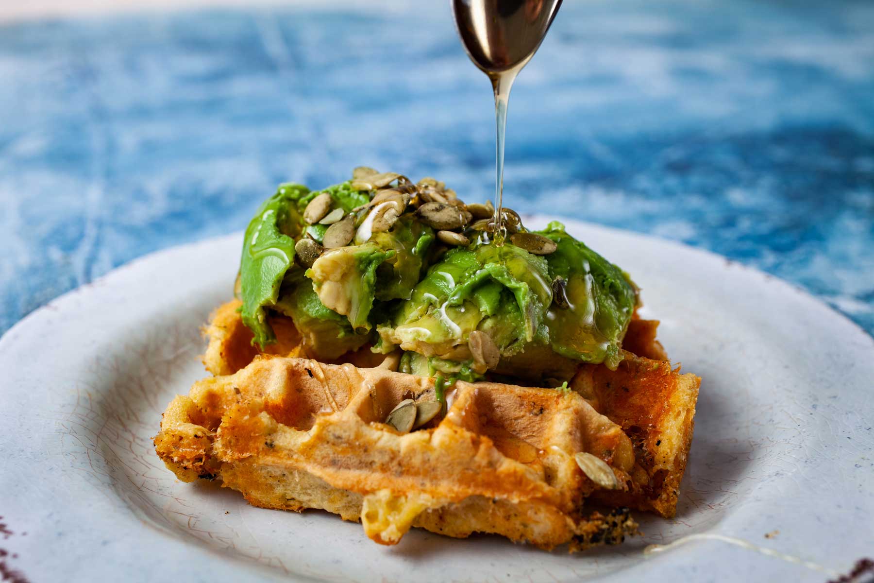 Someone drizzling honey over a waffle topped with avocado.