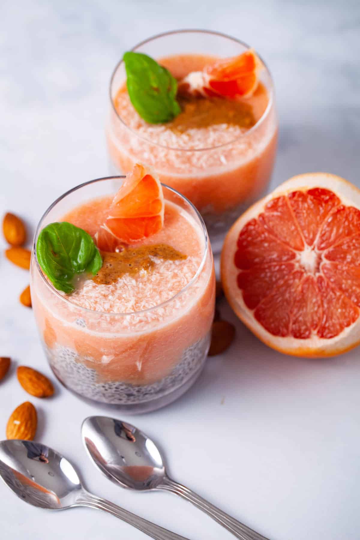 Two glasses filled with chia pudding, infused with a grapefruit sauce, topped with almond butter, Basil leaves, coconut shreds, and slices of grapefruit.