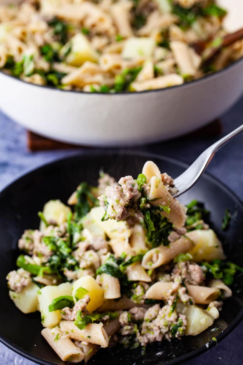 A fork lifting penne with broccoli rabe and sausages over a filled plate.