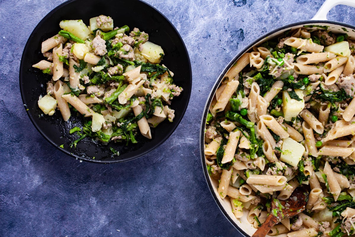 A plate filled with a pasta dish next to a large skillet filled with the same type of food.