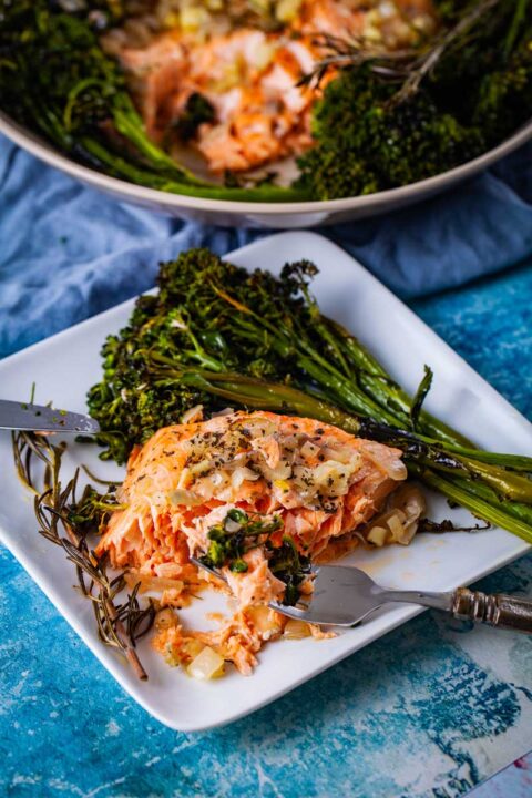 A fork resting on a plate with a piece of baked salmon and a bit of baby broccoli, arranged next to a slab of the pierced in baked salmon and baked broccoli with a sprig of baked thyme on the side.