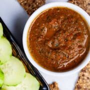 An Italian red pepper spread in a ramekin next to cucumber slices and toast.