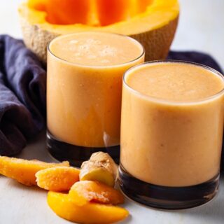 Close-up of two glasses of cantaloupe smoothies shown amongst a few ingredients like half a cantaloupe, more frozen peaches, and ginger.