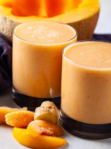 Close-up of two glasses of cantaloupe smoothies shown amongst a few ingredients like half a cantaloupe, more frozen peaches, and ginger.