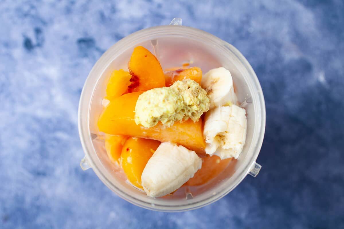 A blender filled with cantaloupe pieces, frozen peaches, pieces of banana, and grated ginger.